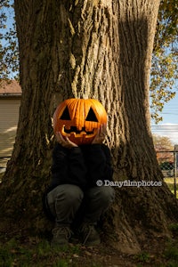 a boy is sitting under a tree with a pumpkin on his face